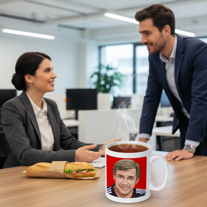 Retrotext 11oz glossy white mug featuring a graphic of Sir Alex Ferguson on a red background, set on an office desk.