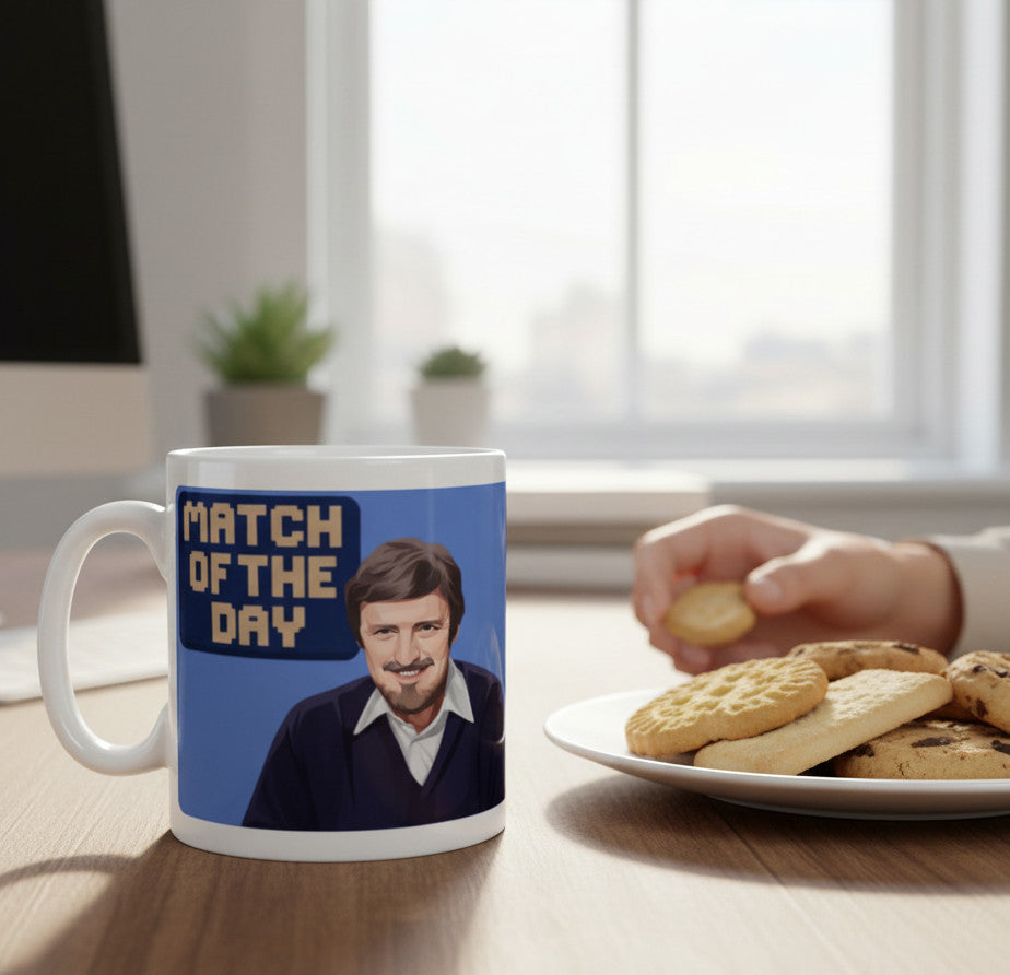 Retrotext 11oz glossy white mug featuring a Jimmy Hill Match of the Day illustration on a desk with a plate of cookies